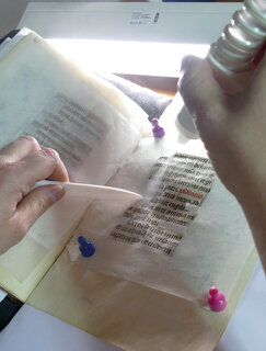 Eine aufgeschlagene Pergamenthandschrift, Teile davon sind abgedeckt. In einem ausgeschnittenen Bereich wird ein Aerosol appliziert, um die Schrift zu fixieren.